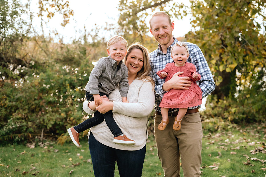 Portrait photograph of Allie (Prion) Gundler '12 smiling as she holds her son in her arms as they are standing next to Allie's husband/son's dad named Zach as Zach is holding their baby daughter in his arms outside somewhere in a grassy lawn area