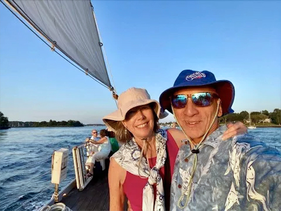 Landscape photograph of Bob Consentino '76 grinning in a faded grey/blue oceanic animal/theme button-up vacation dress shirt and blue sea bucket hat as he poses next to his wife Sharon who is in a dark tan sea bucket hat and a pink tanktop plus white pattern scarf around her neck as they are on a small sailing boat out on the water