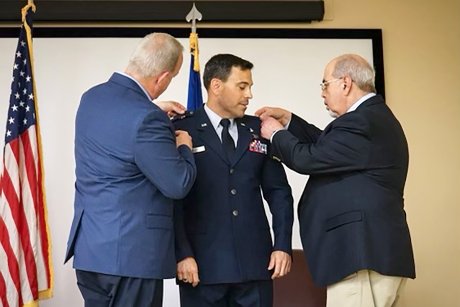 Portrait photograph of Brian Smith '97 in a military outfit suit and tie as he was pinned to LTC in a ceremony in April 2023 as two other men beside him help place small seal ribbon banners on his military suit top in the shoulders area