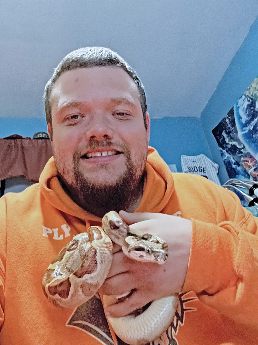 Portrait headshot photograph of Brian Vargas 16' smiling in a orange graphic hoodie while holding his one-year-old Colombian red-tailed boa inside a bedroom area; This snake will grow up to eight feet long! Right now, he is just about three feet long.
