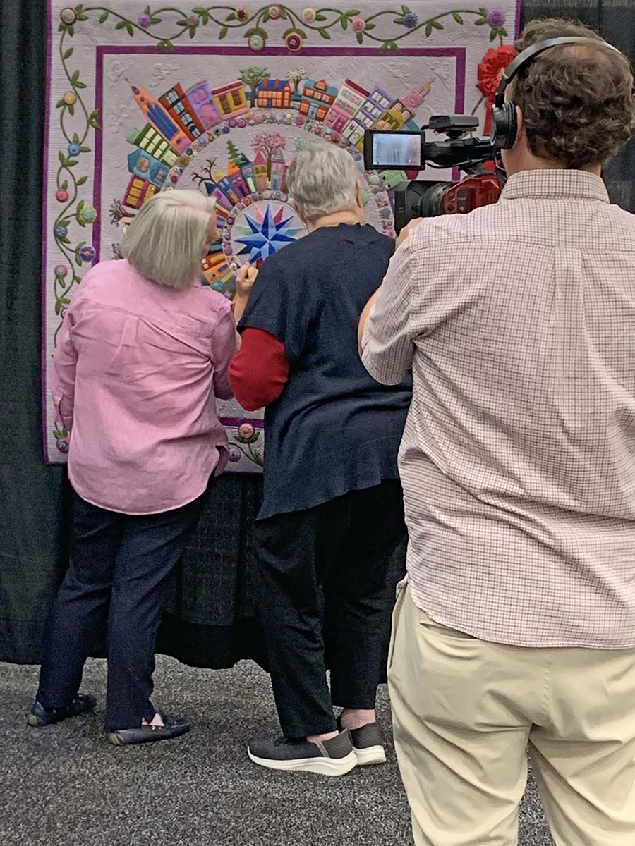 Portrait photograph of Carole (Behan) Berry '79 inside the American Quilters Society show in Grand Rapids, MI as she stands next to another woman beside her in front of Carole's custom-made quilt design pattern (both of them are touching the quilt as they glance at it) while a camera man in a headset and video device is filming behind them