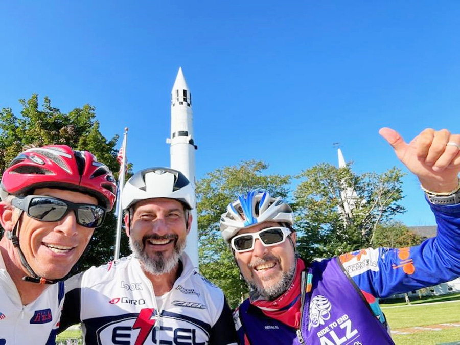 Portrait headshot photograph of David Edry '91, Geoff Hamilton '91, and Peter Stokloza '90 (from left to right) all smiling in bicycling gear and helmets at an Alzheimer's Association fundraising event outside on a bright sunny day