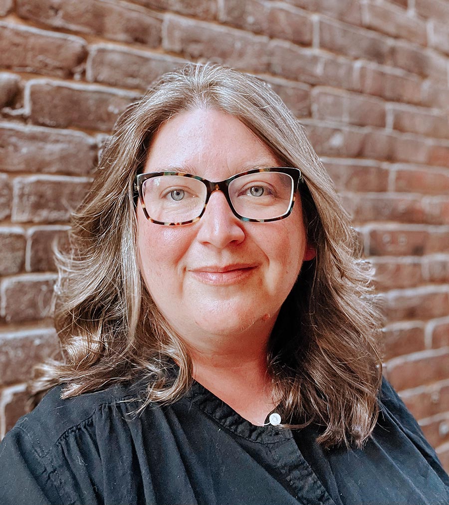 Portrait headshot photograph of Emilie Knisely '07G smiling in a black button-up dress shirt and wearing see through prescription glasses (dark black outer frame/chrome slightly tinted inner frames)