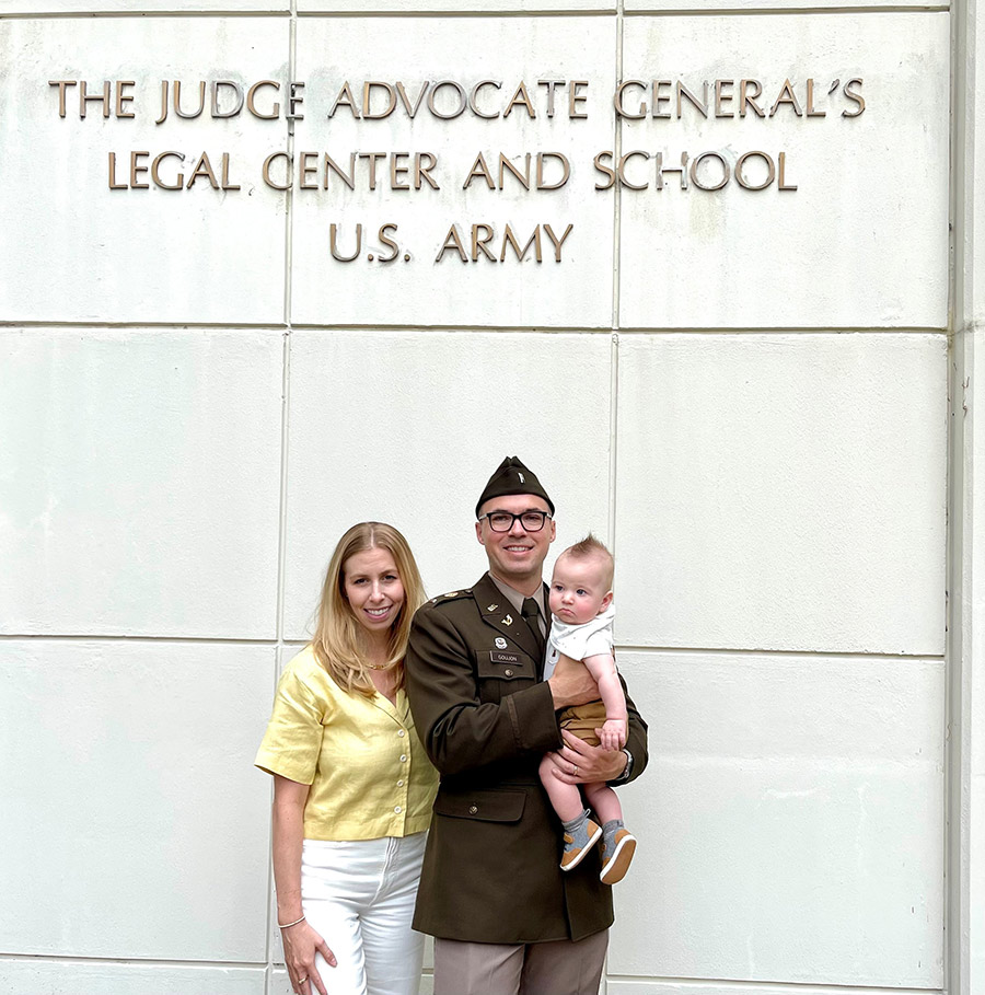 Portrait photograph of First Lieutenant Greg Goujon '10 smiling in his dark brown military suit and cap as he stands next to his wife Sarah and Greg is holding their son Jackson at Greg's graduation in front of The Judge Advocate General's Legal Center and School U.S. Army building display sign located on the grounds of UVA