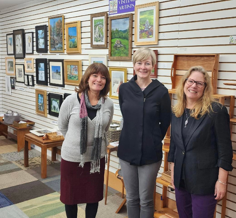 Portrait photograph of Jaylene Bengtson '02G, '08CAGS, Heidi Pettigrew '99, '07G,'11CAGS, and retired PSU Professor of Political Science Sheryl Shirley smiling next to each other inside at the Artistic Roots Visiting Artist gallery room area
