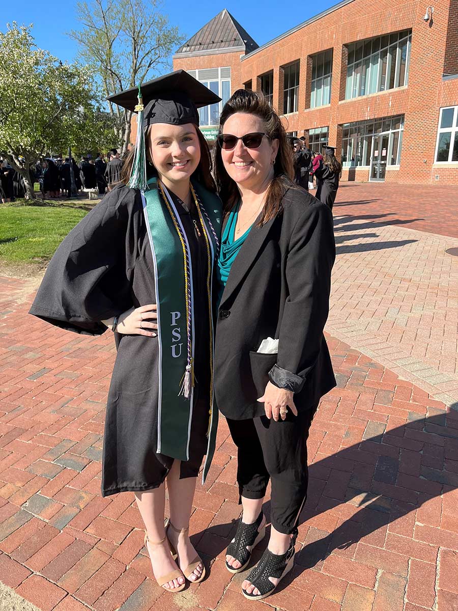Portrait photograph of Holly (Shepherd) King '93 smiling in her graduation gown outfit and cap standing next to Abigail L. King '23 (in a black open coat jacket and turquoise blouse top) on May 13, 2023 at the Plymouth State University Commencement somewhere outside on campus on a bright sunny day