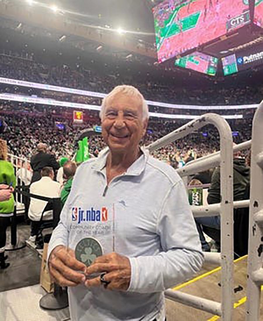 Portrait photograph of Joe Amorosino Sr. '62, smiling in a grey colored open collar jacket as he is holding a Jr. NBA (Celtics) Community Coach of the Year award plaque with the Jr. NBA logo plus Boston Celtics logo shown on the award as he is standing nearby an attached railing guard that is nearby the arena seats inside the TD Garden on March 31, 2023