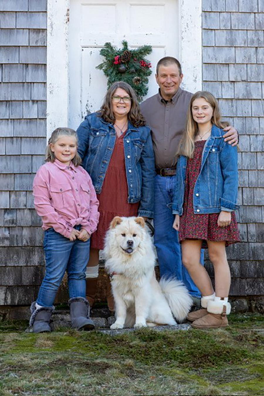 Portrait photograph of Karyn Ames '98 smiling in a dark maroon blouse dress and blue denim jacket standing beside her husband Jeff Polosky and their two daughters Marley and Kiya with the family dog below them as they stand outside their home in Moultonborough