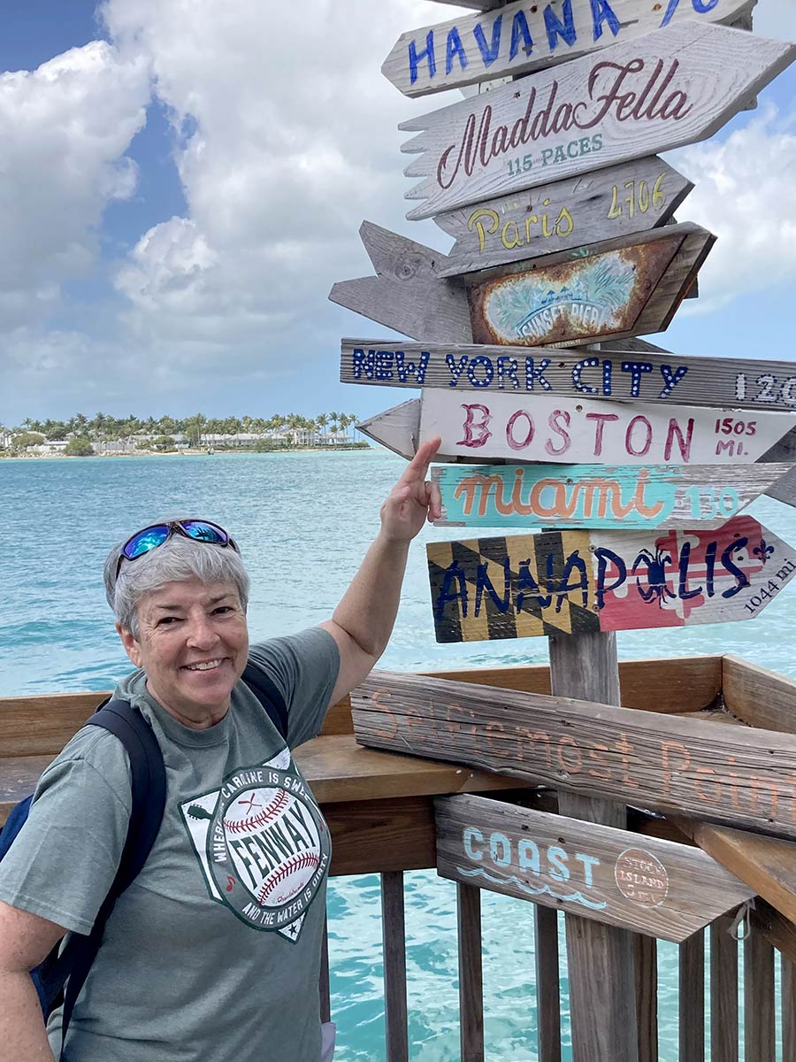 Portrait photograph of Linda Kuhne '82 smiling in a grey Fenway graphic t-shirt, backpack, and sunglasses on her head as she points at a directional wooden post mounted stand that contains few American city names and international locations while her finger is rested on the Boston 1505 mi. display sign as she is standing on a deck area overlooking the ocean in Key West, FL