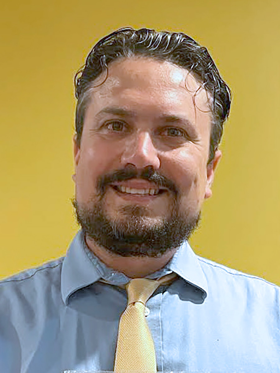 Portrait headshot photograph of Noah Goldblatt '00 smiling in a light sky blue button-up dress shirt and yellow tie