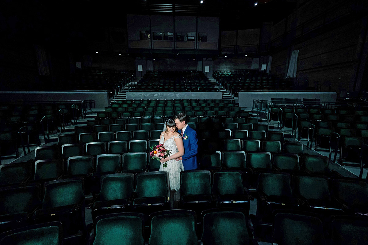 Landscape photograph of Kelly (Rice) Willis ’11 and Jamie Willis ’11 in wedding attire as they pose holding each other smiling inside a dark theater auditorium area setting with Kelly holding a small flower bouquet