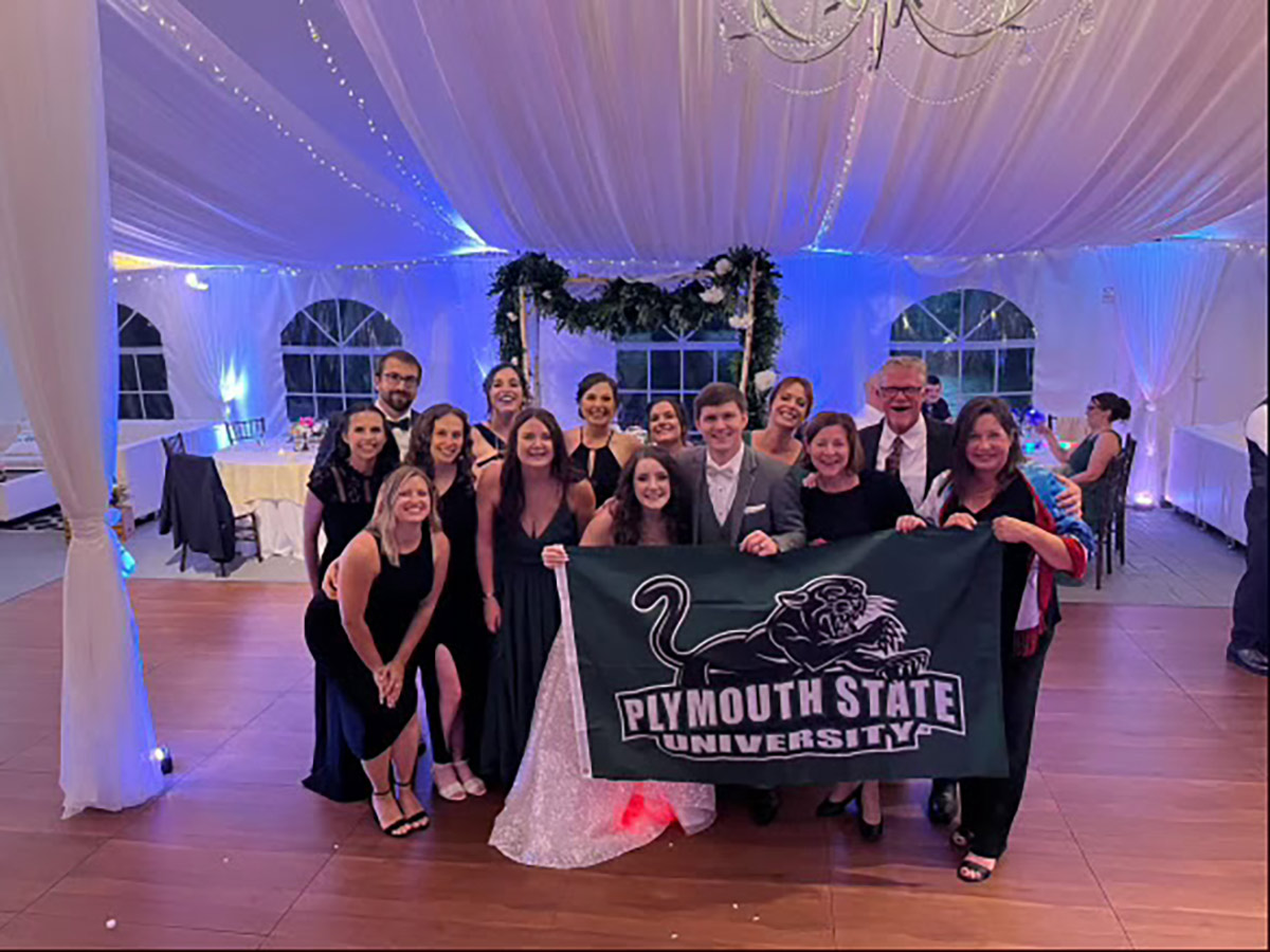 Landscape interior photograph of Ryan Patten '14, '18G and Kate Steinberg's wedding party inside a big white canopy tent area; Individuals featured in the image are [Back row, left to right: Russell Mancini ’15, Hannah Andritsakis ’16, Kayla Gaudette ’14, Forest Steinberg ’14, ’16G, Kimberly Goldberg ’06, Professor Patrick May. Second row, left to right: Tessa Mancini ’14, Danielle Blondin (former assistant director of admissions), Danielle Mishkit (former assistant director of admissions), Ayla Steere ’17, ’19G, Patti May (director of academic support services), Sandy McGarr (associate director of admissions) Center: Kate Patten (bride) and Ryan Patten ’14, ’18G] as the people in the very front row center are holding a Plymouth State University logo flag with school mascot logo above the name