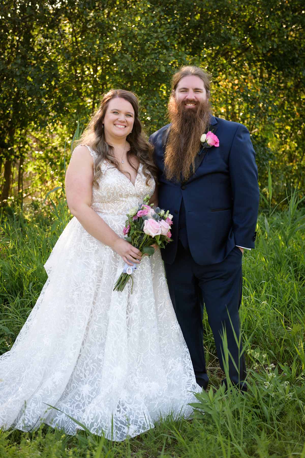Portrait photograph of Chris Rydjeski ’13 and Amberlee Barbagallo ’12 posing outside standing in wedding attire next to each other smiling in a forest grassy area