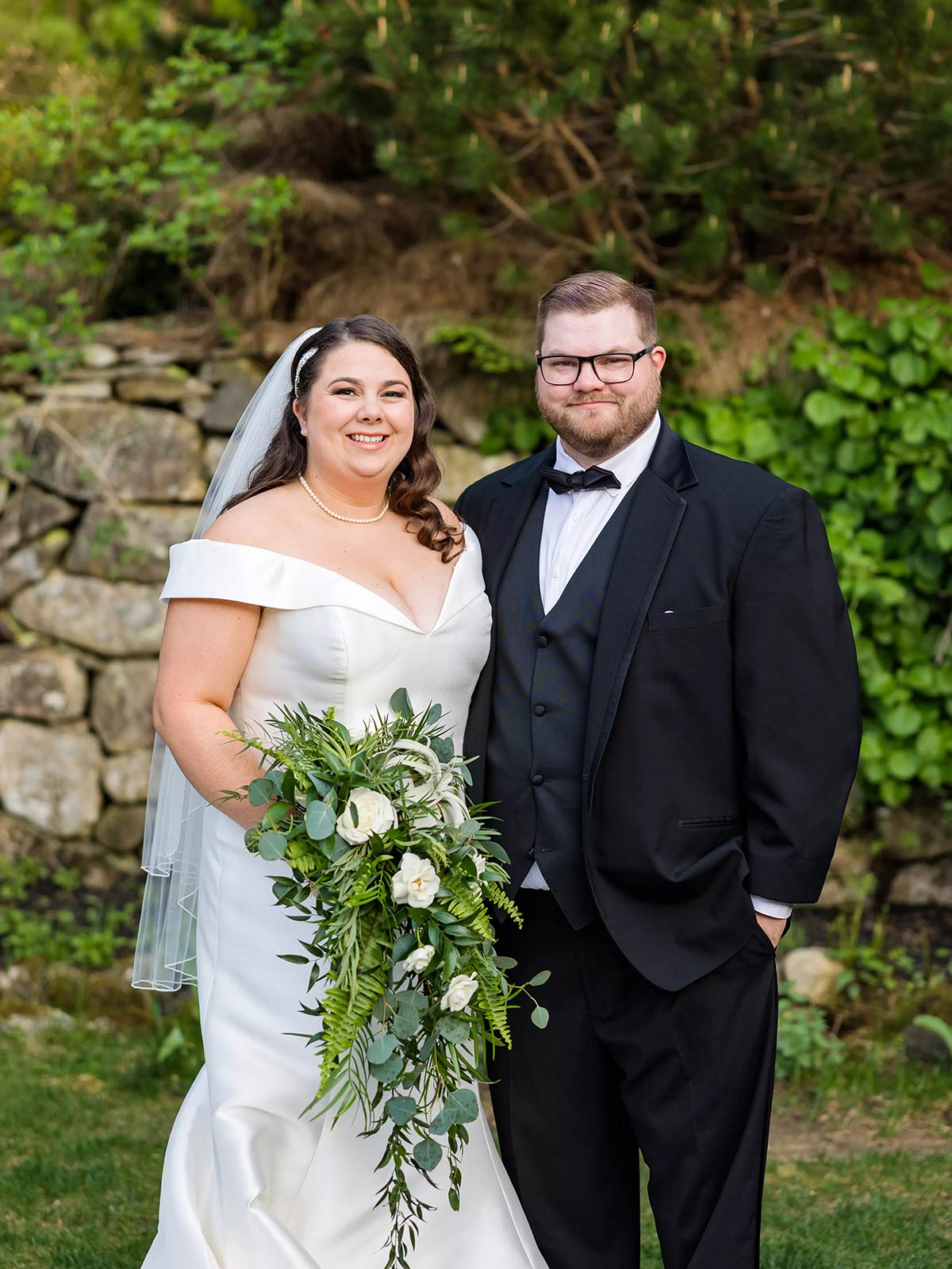 Portrait photograph of Isabelle Elsasser ’19 and Zack Kelley ’19 posing outside standing in wedding attire next to each other smiling in a forest grassy area