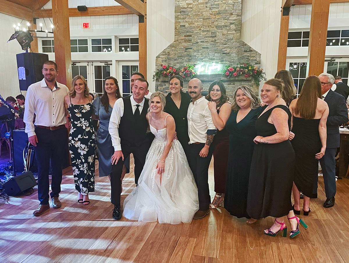 Landscape photograph of Amy Hackett ’17 and Colman Lydon ’17 in their wedding attire posing together smiling in the middle on October 29, 2023 inside on the wooden dance floor at Owl’s Nest Resort; Briana Lumbert ’17 was the officiant. Other PSU alumni present were: Taylor (O’neil) Keith ’16, Ryan Gardent ’17, Casey Henry ’17, Kate McCann ’17, Jeremy D’Amour ’17, Meredith Nicosia ’17, Paige Young ’17, Tucker Moss ’17, and Kaitlyn Warren ’15.