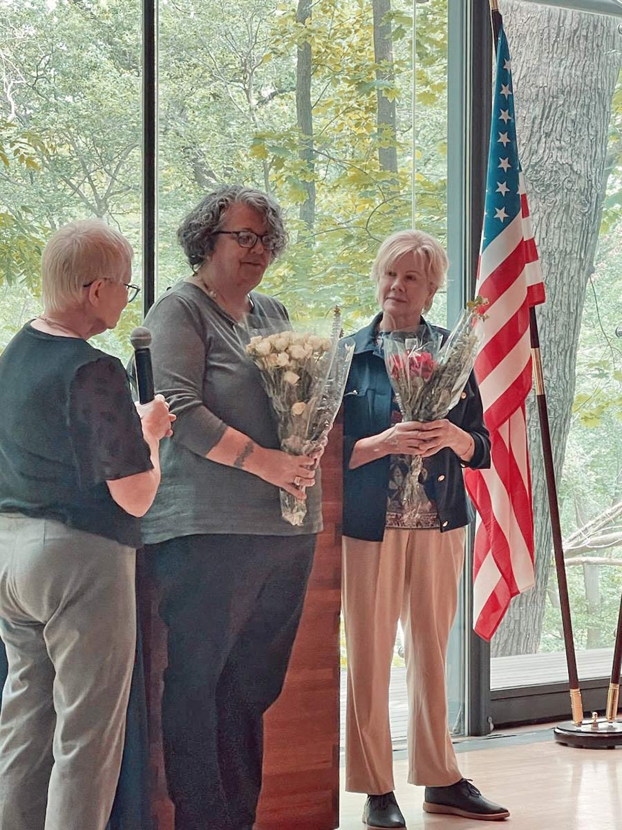 Portrait photograph of Rachel Simoneau-Mueller ’98 and Kaija Wilson holding their own small flower bouquets at the Finnish Embassy in DC accepting their presidential roles for Finn Spark Kipinäkerho while a presenter speaker woman is holding a microphone to congratulate them