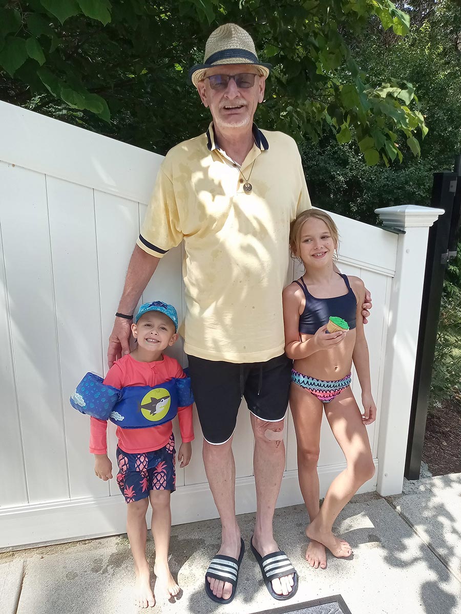 Portrait headshot photograph of Reiner Bertelmann '73 smiling in a yellow beach polo top and dark tan colored trilby hat with sandals standing next to his two precious grandchildren as they are wearing beach swimwear as they are all outside standing next to the edge of a white jagged fence