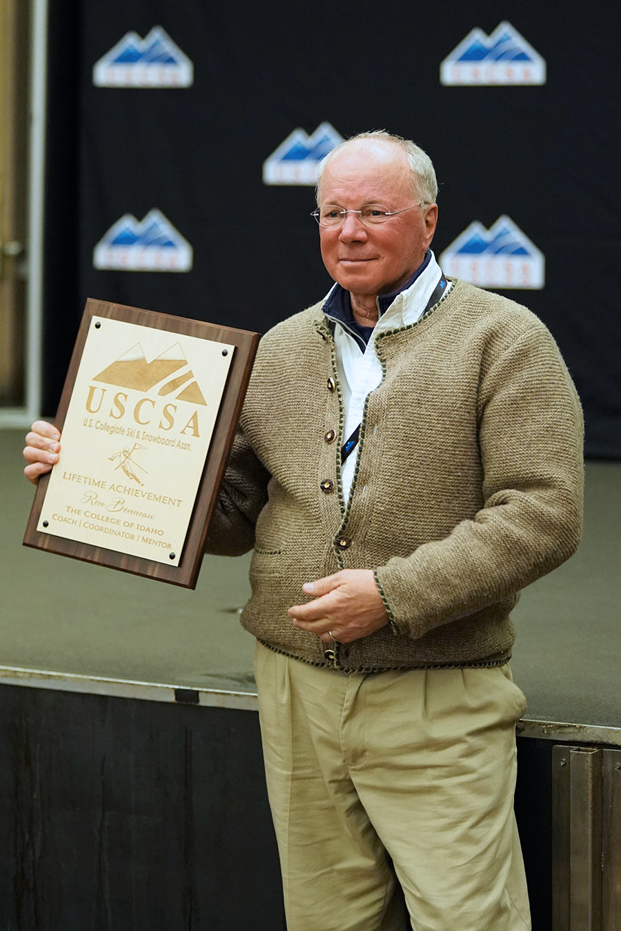 Portrait photograph of Ron Bonneau ’81 grinning in a dark tan wooly cardigan jacket as he holds the Lifetime Achievement award plaque in his right hand from the US Collegiate Ski & Snowboard Association at the USCSA National Championship awards ceremony in Mammoth Lakes, CA