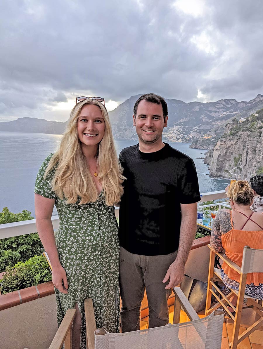 Portrait photograph of Stefan Slattery '13 in a black shirt/dark brown jeans & Louise Smith in a green floral dress with sunglasses on top of her head in Praiano, Italy smiling as they are standing next to each other on a rooftop patio edge area overlooking the mountain topography and ocean
