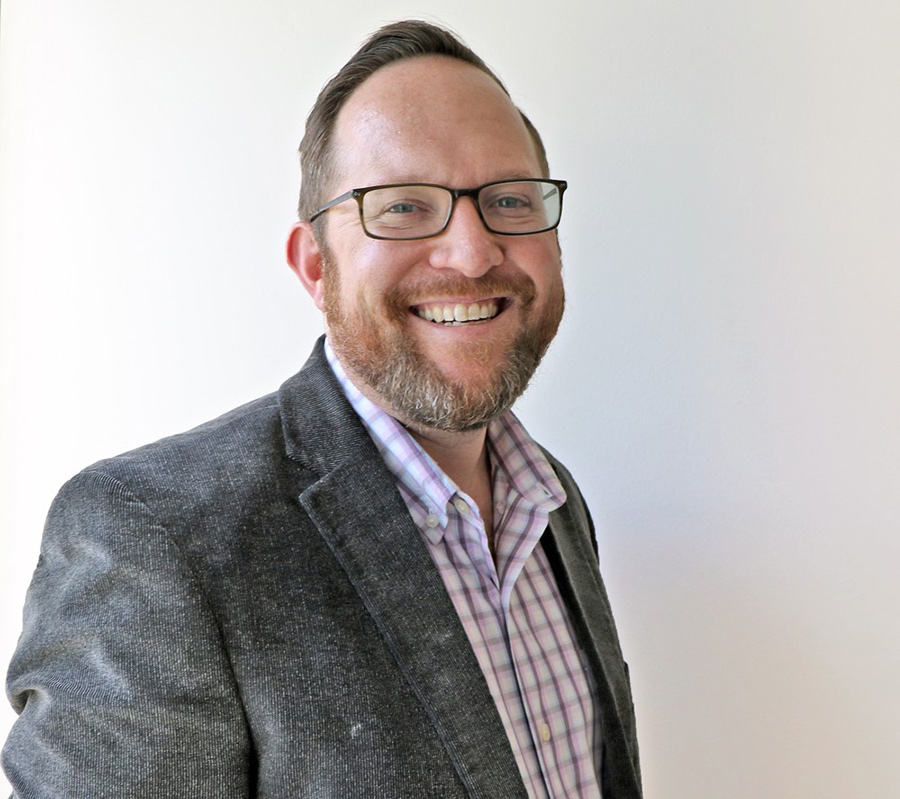 Portrait headshot photograph of Timothy Carrigan '04 smiling in a multi-colored button-up dress shirt (purple/white) and open charcoal grey colored business coat jacket plus has on prescription glasses (dark black outer frame/chrome slightly tinted inner frames)