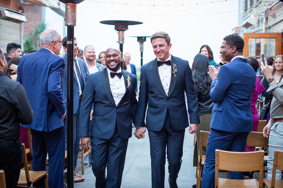 Portrait photograph of Trevor Chandler '09 smiling in a dark navy blue suit and black bowtie holding hands with his significant romantic partner (a man) with the happy partner wearing the same outfit as Trevor where both walk down an aisle outside at a wedding event as many other people celebrate nearby them