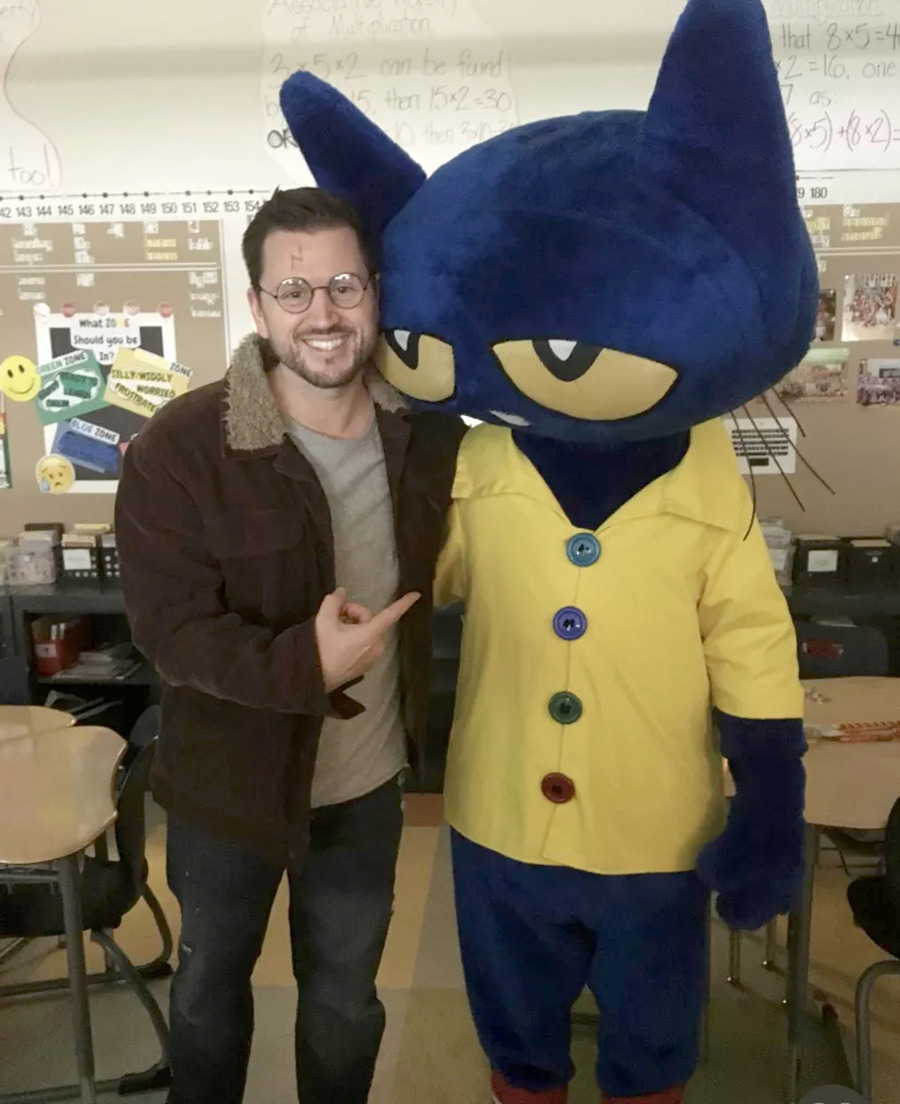 Portrait photograph of Zachary Cary '04 smiling in a Harry Potter character style outfit and pointing at Pete the Cat blue mascot in a yellow button-up shirt in Zachary's classroom area