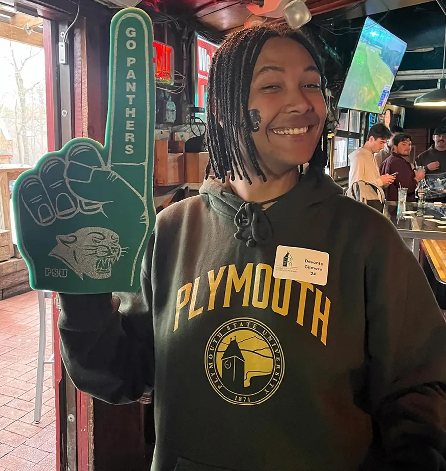 a male student wearing a Plymouth sweatshirt and a PSU foam finger smiles brightly while attending the Pints with PSU event at the Lucky Dog Tavern