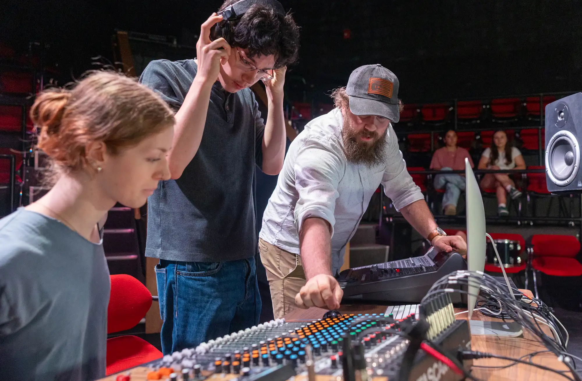 Recording Studio Supervisor Parish Dawe-Chadwick, in a light blue dotted shirt and cap, adjusts a sound mixing console while a Plymouth student wearing headphones looks on and another Plymouth student observes in the foreground