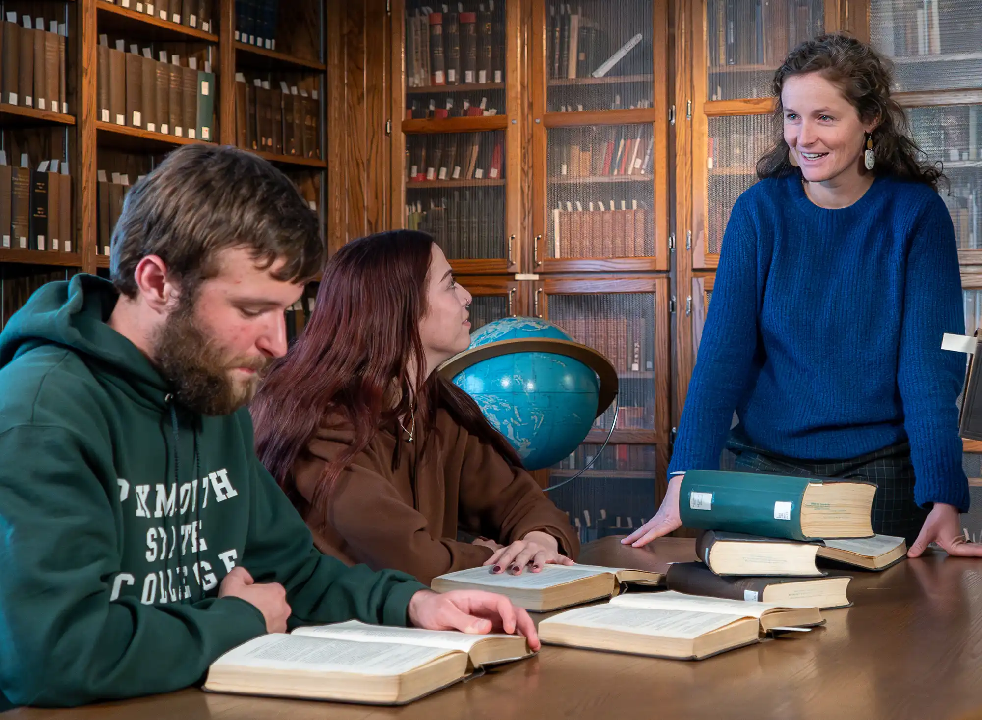 Professor Kelsie Eckert stands by a table in a library, looking at two Plymouth University students who are seated with open books.
