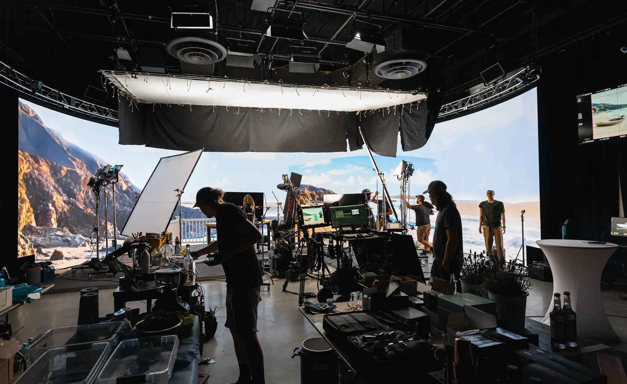 A busy virtual production set with a large curved LED screen displaying a coastal landscape, numerous crew members, cameras, lighting rigs, and production equipment.