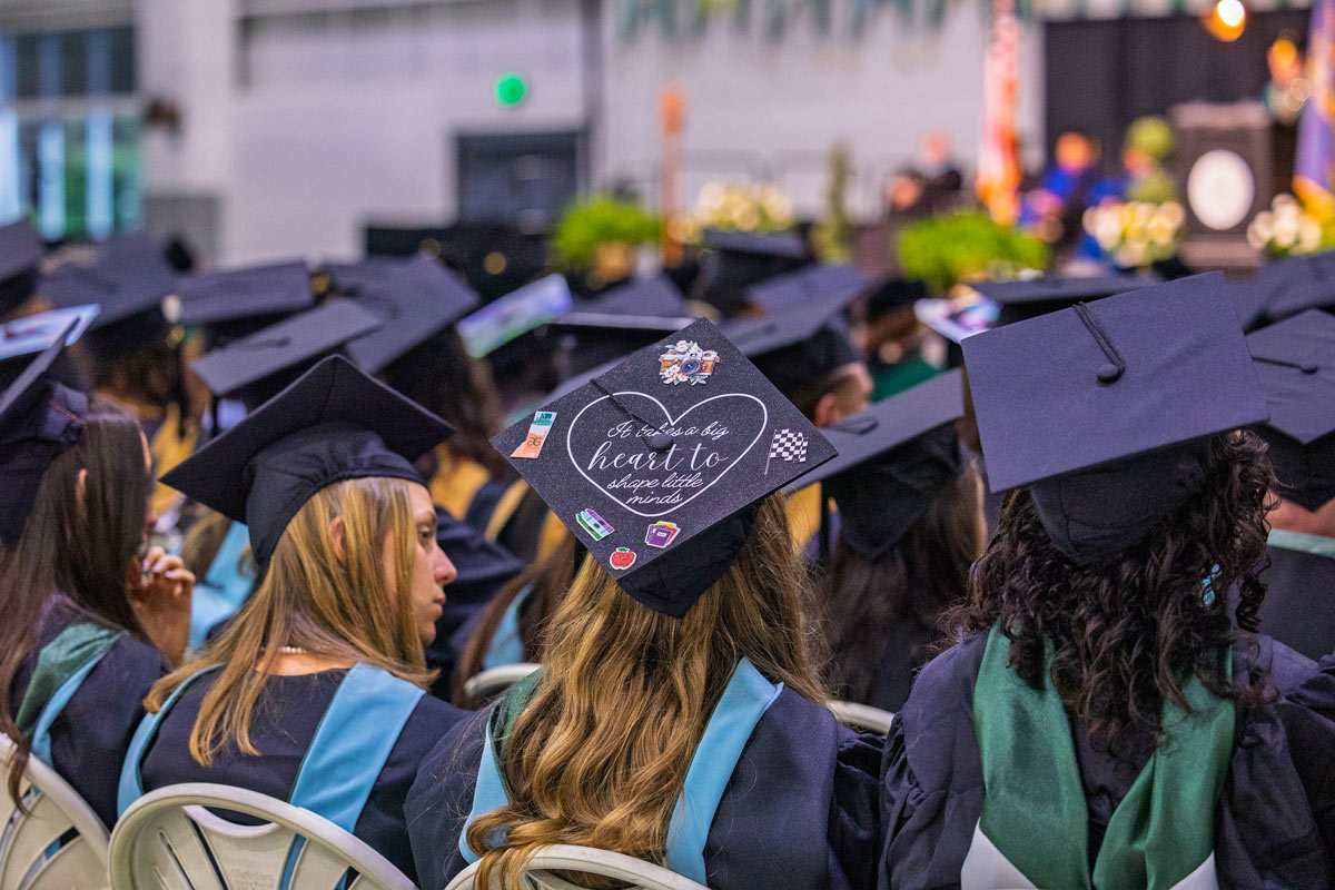 A rear-facing medium shot captures a group of graduates seated in a hall, focusing on their graduation caps. In the foreground, a cap is decorated with white text that reads, "It takes a big heart to shape little minds," surrounded by small illustrations of school supplies, a heart, and a checkered flag.