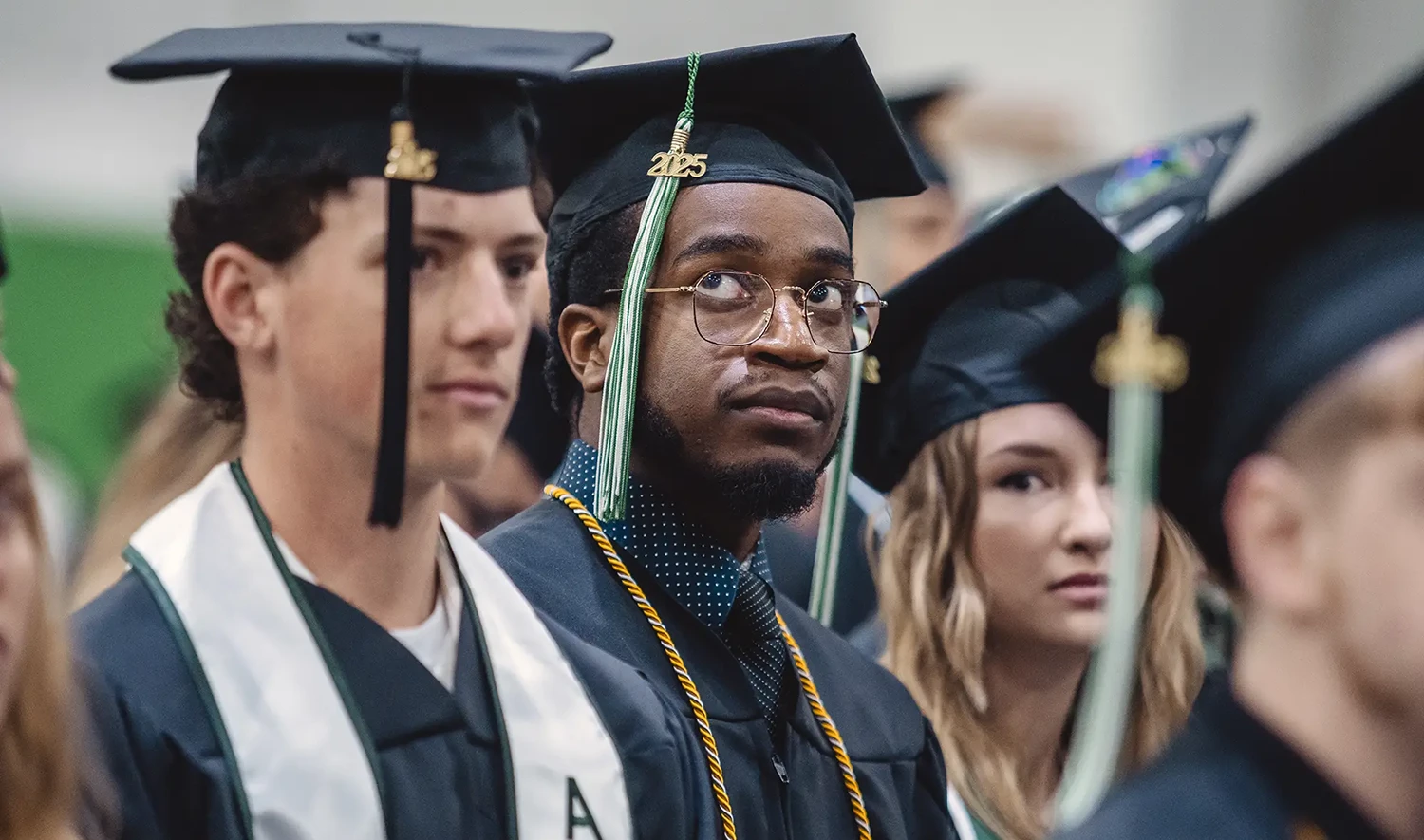 A medium shot of three young adults at a graduation ceremony. In the foreground, centered, is a person wearing glasses and a graduation cap with "2025" on it, looking intently to the right.