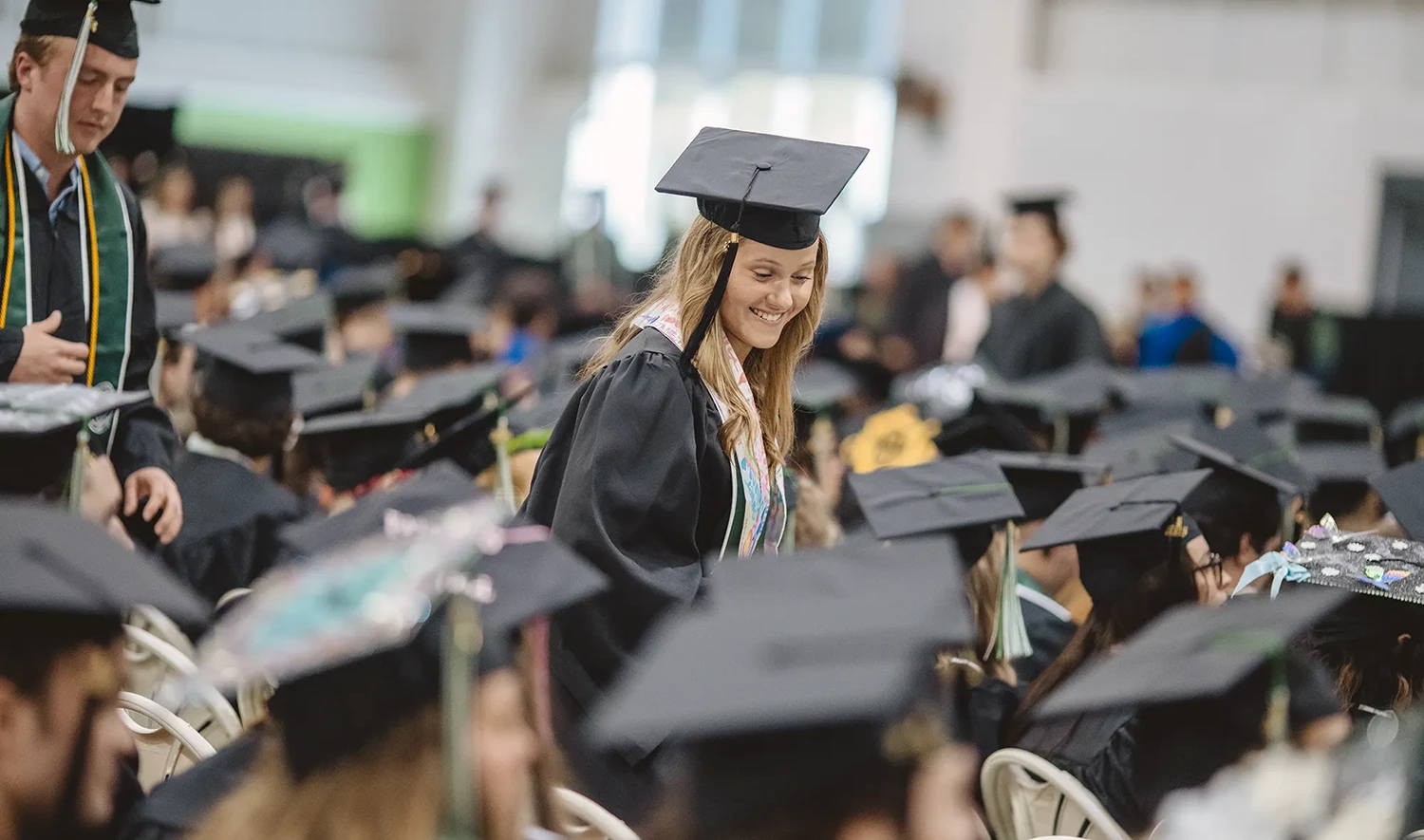 A candid shot captures a young woman in the center of the frame, smiling and looking down, surrounded by other graduates.