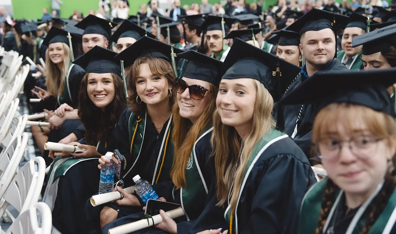 A row of graduates are seated in white folding chairs during a commencement ceremony. The person in the foreground on the right is partially visible, wearing glasses and a graduation cap. From left to right, the next four individuals are smiling at the camera.